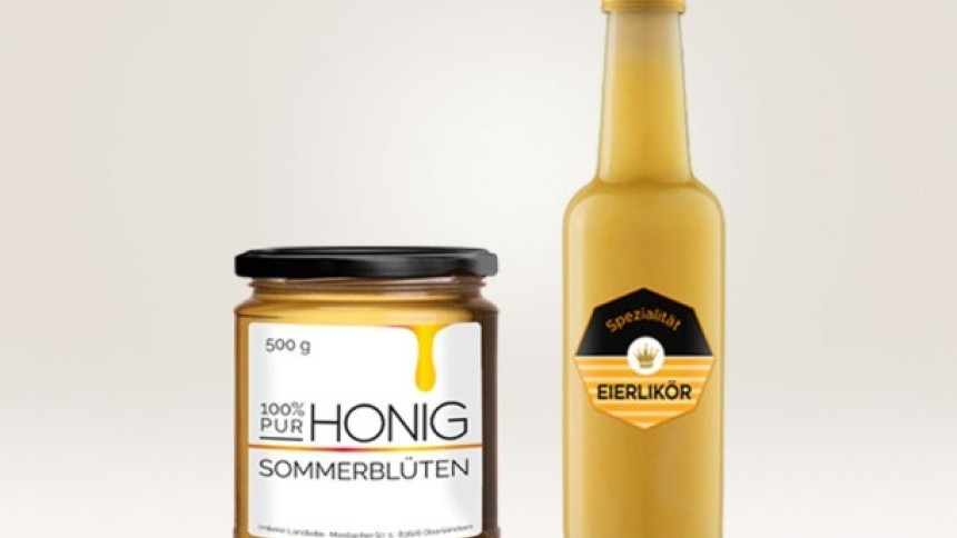 Staklenka meda s bijelom etiketom i crnim čepom stoji pokraj visoke boce kremastog likera od jaja s žuto-crnom etiketom na svijetloj pozadini.