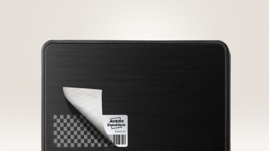 Crna pravokutna površina s zaobljenim kutovima, s malom srebrnom sigurnosnom naljepnicom zalijepljenom u donjem lijevom kutu. Kut naljepnice je djelomično odljepljen, otkrivajući ispod šahovnicu; vidljivi dio naljepnice prikazuje logotip "Avery Zweckform" i mali crtični kod.