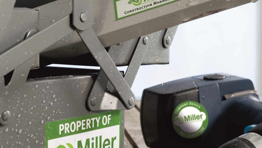 Sivi metalni građevinski uređaj s vidljivim vijcima i kapljicama vode. Na konstrukciji su pričvršćene zelene i bijele oznake vlasništva s logotipom Miller i tekstom 'PROPERTY OF Miller'; slična okrugla oznaka nalazi se na crnom električnom alatu pored njega.