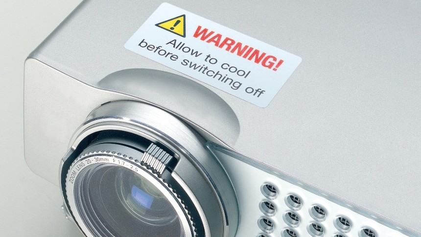 Srebrni projektor s velikim objektivom sprijeda. Na kućištu se nalazi bijela naljepnica za upozorenje s natpisom „WARNING! Allow to cool before switching off” i trokut sa simbolom upozorenja.