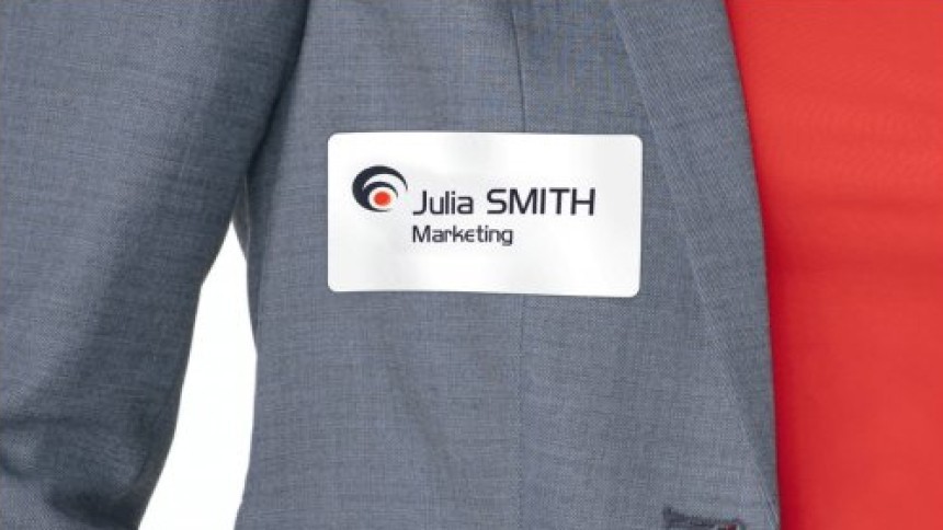 Bijela identifikacijska oznaka s otisnutim logotipom tvrtke, imenom i odjelom „Marketing” pričvršćena je na sivoj jakni preko crvene košulje.