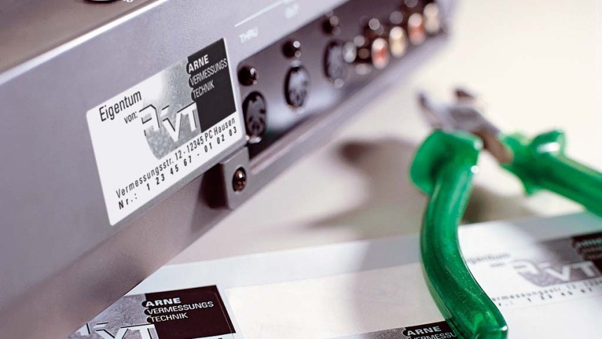 Srebrna pločica s logotipom tvrtke i natpisom zalijepljena je na kućište elektroničkog uređaja; pored nje su zelene kliješta i druga pločica.