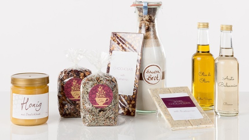Skupina različitih prehrambenih proizvoda s elegantnim etiketama. Na fotografiji se vide staklenka meda, dva pakiranja čaja, čokolada, mješavina za pečenje kruha, bijela čokolada te boce maslinovog ulja i octa.