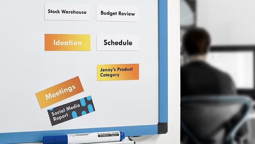 Zbliżenie na białą tablicę magnetyczną w niebieskiej ramie. Na tablicy przyczepione są drukowane etykiety z napisami: "Stock Warehouse", "Budget Review", "Ideation", "Schedule", "Jenny's Product Category", "Meetings" oraz "Social Media Report". Na dolnej półce leży niebieski marker.