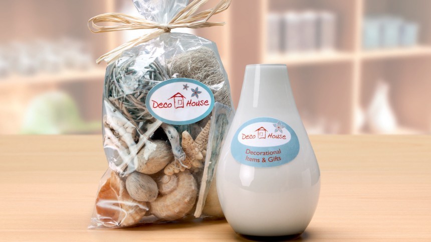 Poklon set: prozirna vrećica sa školjkama i bijela vaza. Oba proizvoda ukrašena su ovalnim naljepnicama s logotipom "Deco House", prikazujući dosljedno brendiranje za dekorativne predmete.