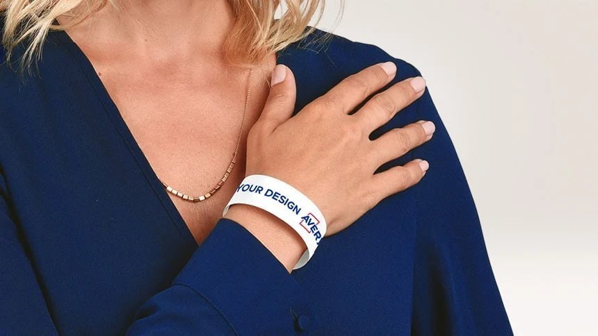 Žena u plavoj bluzi nosi na zapešću bijelu narukvicu s natpisom "YOUR DESIGN HERE", prikazujući mogućnost personalizacije proizvoda.