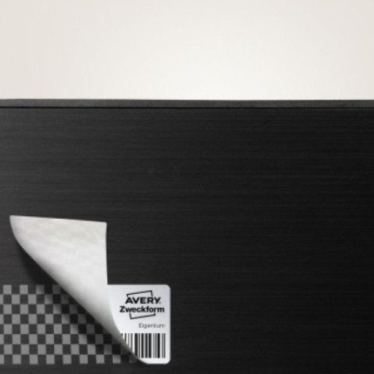 Crna pravokutna površina s zaobljenim kutovima, s malom srebrnom sigurnosnom naljepnicom zalijepljenom u donjem lijevom kutu. Kut naljepnice je djelomično odljepljen, otkrivajući ispod šahovnicu; vidljivi dio naljepnice prikazuje logotip "Avery Zweckform" i mali crtični kod.