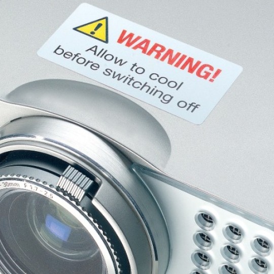 Srebrni projektor s velikim objektivom sprijeda. Na kućištu se nalazi bijela naljepnica za upozorenje s natpisom „WARNING! Allow to cool before switching off” i trokut sa simbolom upozorenja.