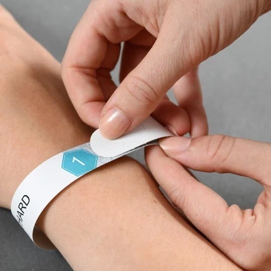 Osoba stavlja bijelu identifikacijsku narukvicu oko zapešća, odljepljujući zaštitnu traku sa samoljepljivog kraja.