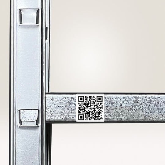 Dio metalne skladišne police s malom, bijelom naljepnicom s QR kodom zalijepljenom na vodoravnu gredu, namijenjenom za skeniranje lokacije ili proizvoda.