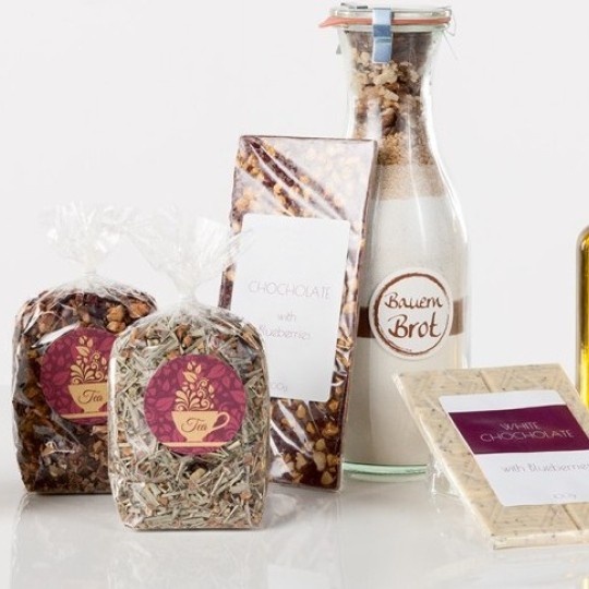 Skupina različitih prehrambenih proizvoda s elegantnim etiketama. Na fotografiji se vide staklenka meda, dva pakiranja čaja, čokolada, mješavina za pečenje kruha, bijela čokolada te boce maslinovog ulja i octa.