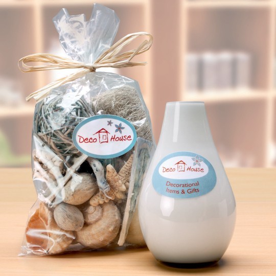Bijela keramička vaza i prozirna vrećica sa školjkama i drugim ukrasima. Oba proizvoda imaju etiketu s logotipom "Deco House".