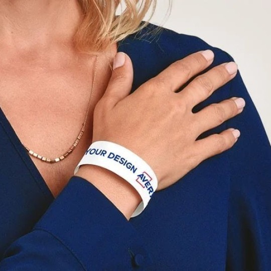 Žena u plavoj bluzi nosi na zapešću bijelu narukvicu s natpisom "YOUR DESIGN HERE", prikazujući mogućnost personalizacije proizvoda.