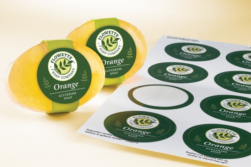 Dva okrugla, narančasta glicerinska sapuna sa zelenim naljepnicama "Flowette Soap Company". Pored njih leži list s istim ovalnim naljepnicama za samostalno ispisivanje, ilustrirajući proces etiketiranja proizvoda.