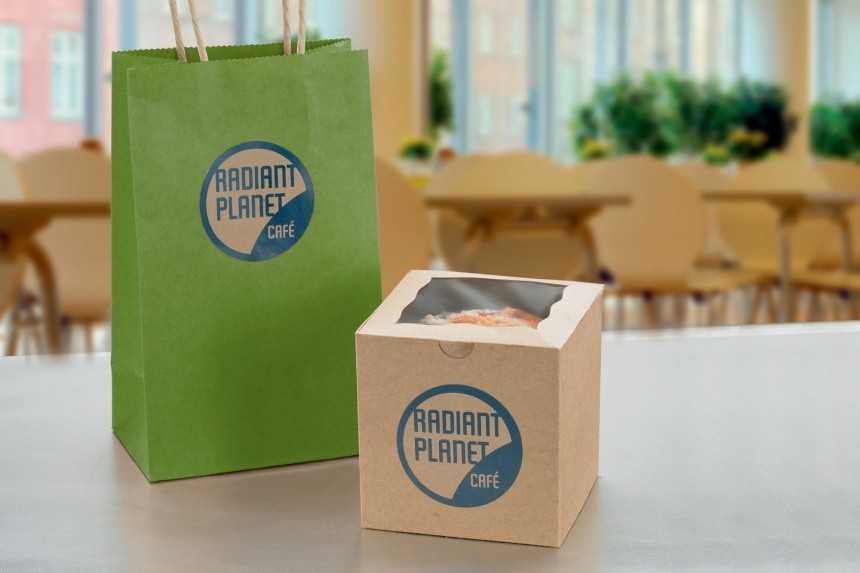 Zelena papirnata vrećica i kartonska kutija za kolač, oboje s okruglom naljepnicom s logotipom "Radiant Planet Café". Komplet prikazuje primjer brendiranja ambalaže za van za kafić.