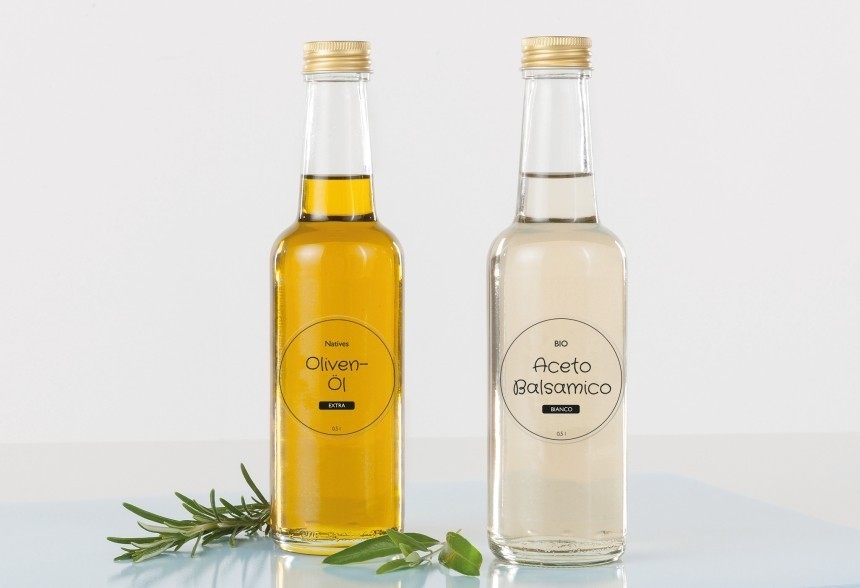 Dvije staklene boce: jedna s ekstra djevičanskim maslinovim uljem, a druga s bijelim balzamičnim octom. Ispred njih leži grančica svježeg ružmarina. Etikete su okrugle i minimalističke.