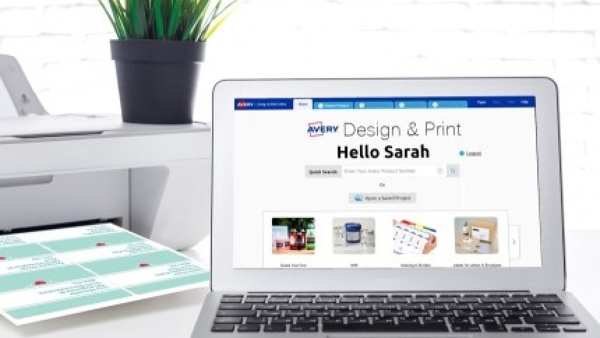 Otvoreni laptop s programom "Avery Design & Print" na zaslonu, a pored njega pisač koji ispisuje list personaliziranih etiketa.