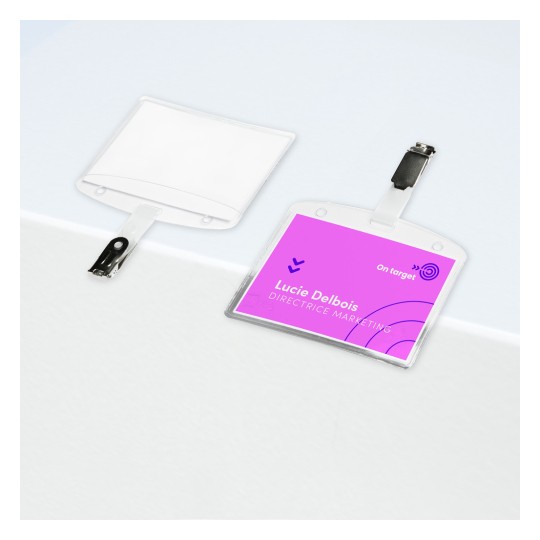 Slika prikazuje dvije pločice s imenom, jednu bijelu i praznu, a drugu s ružičastom pozadinom na kojoj su ispisani ime 'Lucie Delbois' i naziv 'DIREKTORICA MARKETINGA'. Obje pločice imaju kopču za pričvršćivanje.