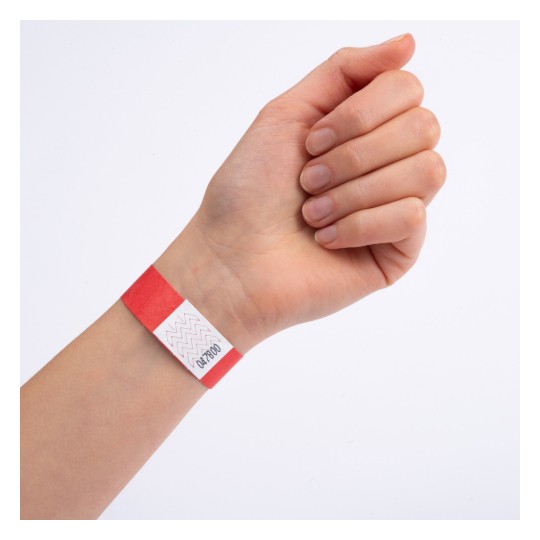 Ruka koja nosi crvenu narukvicu s bijelom etiketom na kojoj su otisnuti cik-cak uzorci i brojevi.