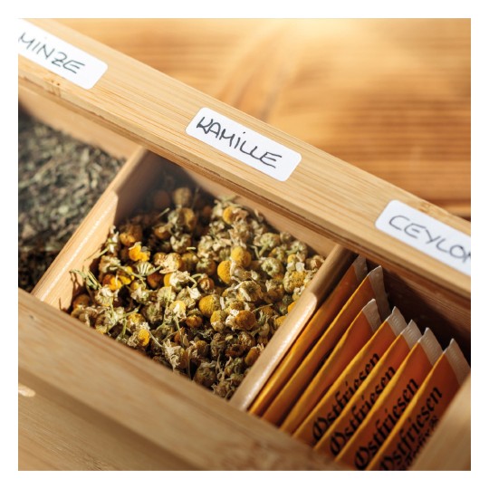 Slika prikazuje drvenu kutiju koja sadrži razne biljne čajeve. Prikazuje pregrade označene različitim vrstama čajeva, uključujući kamilicu (kamille) i cejlonski čaj, s vidljivim suhim cvjetovima kamilice u jednom odjeljku.