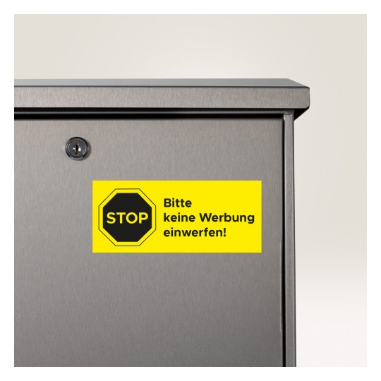 Slika prikazuje krupni plan poštanskog sandučića s jarko žutom naljepnicom na kojoj piše "STOP bitte keine Werbung einwerfen!", što znači da se reklamni materijali ne bi trebali ubacivati.