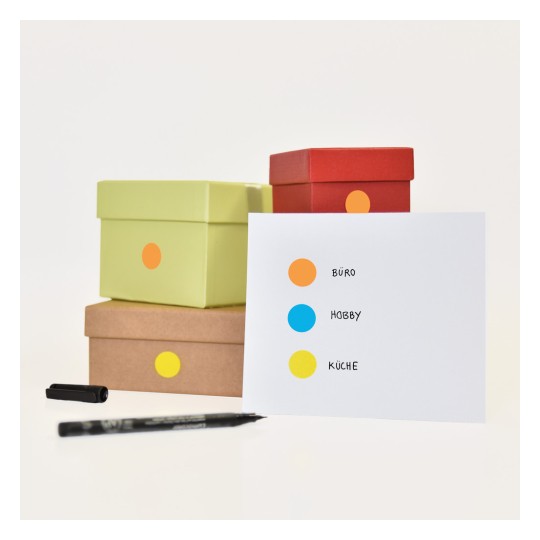 Slika prikazuje nekoliko šarenih kutija naslaganih zajedno s karticom koja ima tri označene točke: narančasta za 'BÜRO', plava za 'HOBBY' i žuta za 'KÜCHE'. Crna olovka također je vidljiva pored kutija.