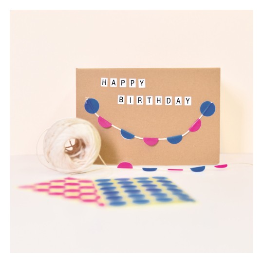 Slika prikazuje rođendansku čestitku s ispisanim riječima 'SRETAN ROĐENDAN'. Na kartici je ukrasna traka s plavim i ružičastim krugovima. U blizini se nalazi kolut špage i uzorak papira s raznobojnim točkama.