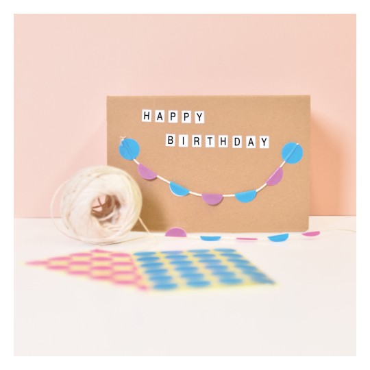 Slika prikazuje smeđu karticu s natpisom 'SRETAN ROĐENDAN' ispisanim slovima. Preko kartice visi ukrasna girlanda napravljena od plavih i ljubičastih krugova. U prvom planu nalazi se kolut konopa, zajedno s uzorkovanim papirom u ružičastoj, žutoj i plavoj boji.