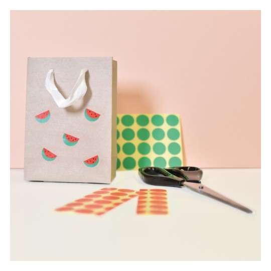 Slika prikazuje ukrasnu papirnatu poklon vrećicu ukrašenu naljepnicama s motivom lubenice. Uz nju se nalazi papir s uzorkom, par škara i šarene okrugle naljepnice raspoređene na ravnoj površini s nježno ružičastom pozadinom.