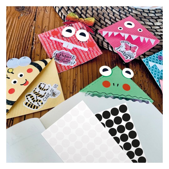 Slika prikazuje kolekciju šarenih, maštovitih dizajna omotnica koje nalikuju raznim likovima, uključujući čudovište, pčelu i žabu. Postavljeni su na drvenu površinu uz uzorirani papir i prazan bilježnicu.