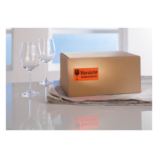 Slika prikazuje kartonsku kutiju s upozorenjem 'Vorsicht zerbrechlich!' što znači 'Opasnost! Lomljivo!' pored dvije čaše za vino na stolu.