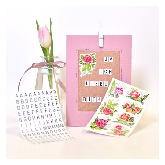 Slika prikazuje ružičastu karticu s tekstom 'JA ICH LIEBE DICH' (volim te), ukrasne naljepnice s cvijetovima, malu vazu s tulipanom i list s naljepnicama abecede.