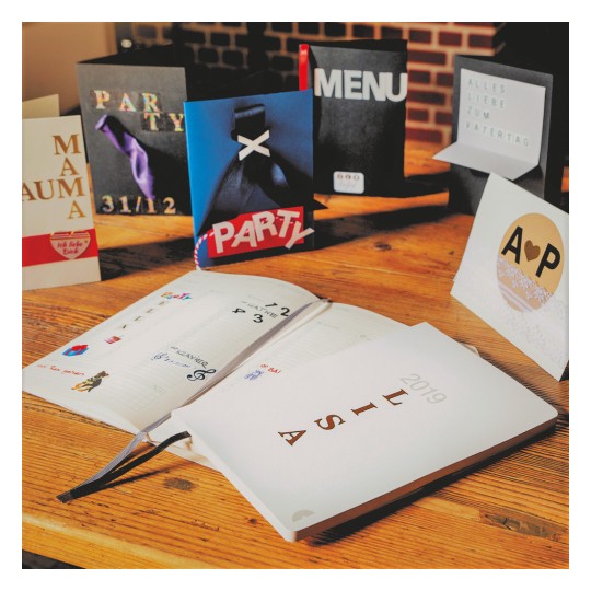 Slika prikazuje kolekciju čestitki i planer na drvenom stolu. Čestitke uključuju teme kao što su pozivnice za zabavu i jelovnici, dok je planer otvoren i prikazuje različite unose i ukrase.