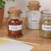 Slika prikazuje tri staklene staklenke sa začinima: jedna s oznakom 'Chili' sadrži sušene čili paprike, druga s oznakom 'Salz' ispunjena je solju, a treća s oznakom 'Oregano' sadrži suhi origano. Tu je također i lončanica za biljke te prazne bilješke s drvenim štapićem na drvenoj površini.